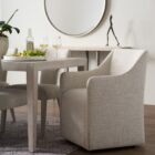 Contemporary upholstered dining chair in soft neutral fabric with clean lines, low arms, and a fully skirted base.