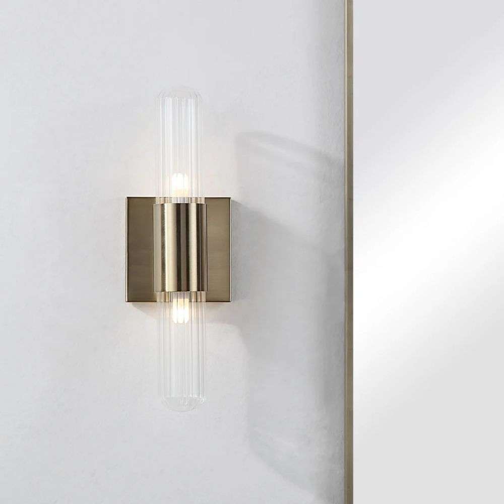 Modern wall sconce with brass backplate and vertical ribbed glass light tubes creating a soft ambient glow.