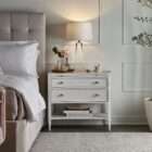 White nightstand with two drawers, slim legs, brass-accented hardware, and an open lower shelf.