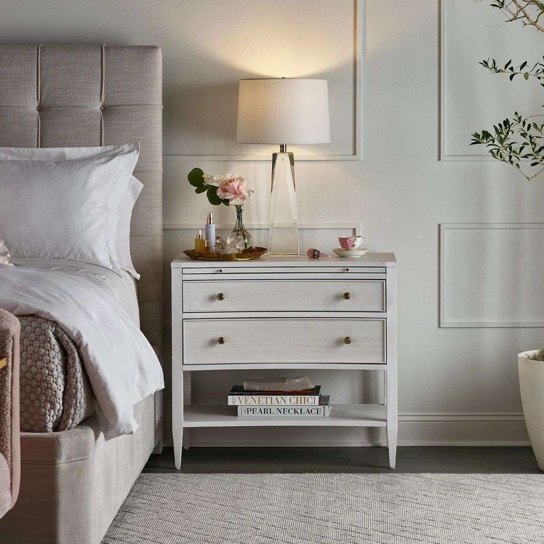 White nightstand with two drawers, slim legs, brass-accented hardware, and an open lower shelf.