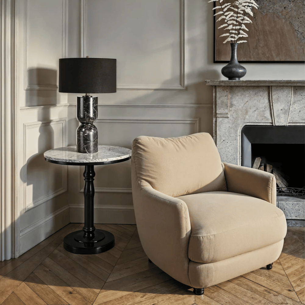 Black marble table lamp styled on a round side table beside a cream armchair and stone fireplace.